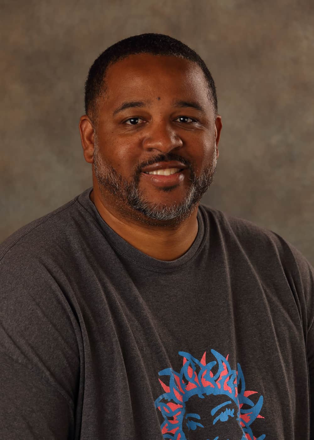 Photo of Lead Teacher Alan Powe, He is an African-American man with short hair and a beard. He is smiling and wears a dark gray shirt with a pink and blue picture of the Sun on it.
