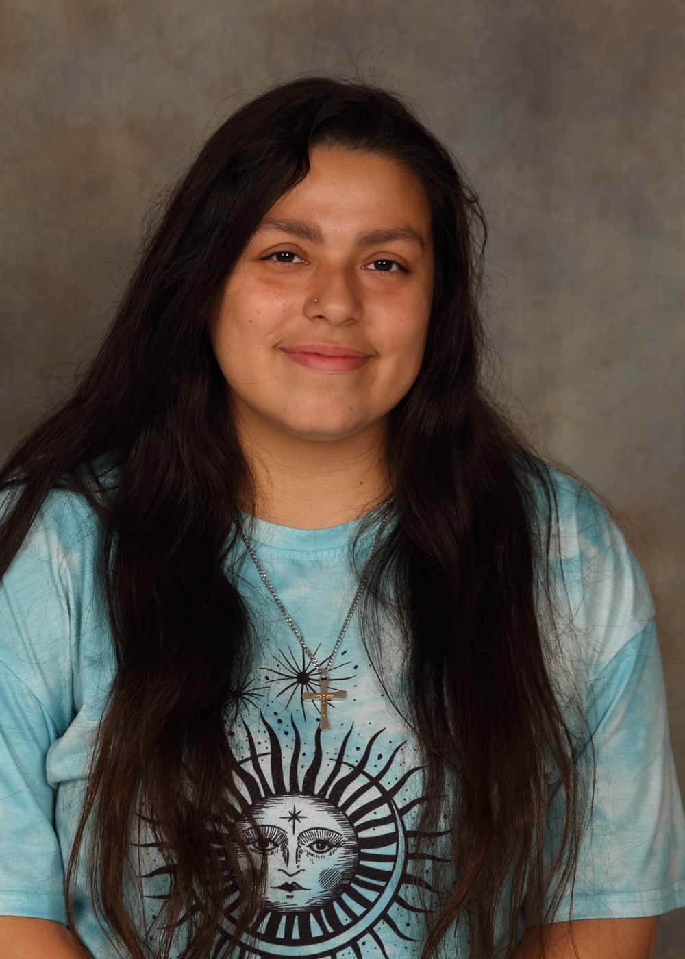 Photo of Co-Teacher Amy Chech. She is a Caucasian woman with long dark brown hair and brown eyes. She is smiling and wears a gold cross necklace and light blue ti-dyed T-shirt with a stylized black drawing of the Sun on it.