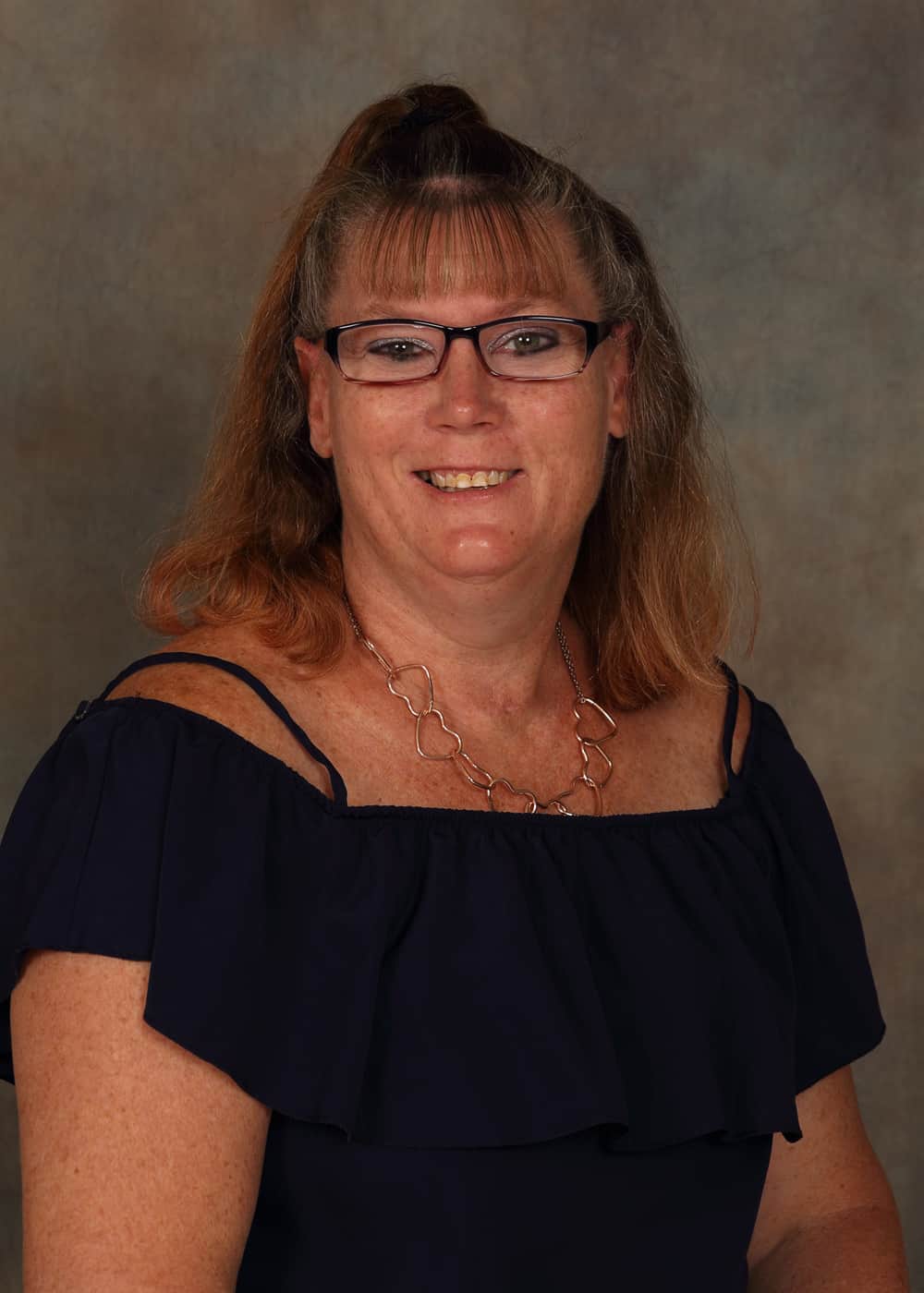 Photo of Lead Teacher Candi Heitkam. She is a Caucasian woman with long brown hair, dark-framed glasses and blue eyes. She is smiling and wears a gold necklace made from interlaced hearts and a navy blue dress.
