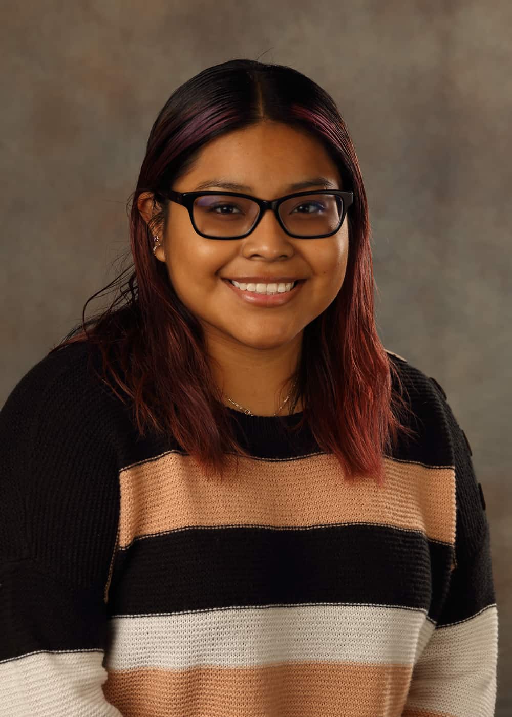 Photo of Co-Teacher Cathleen Escamilla. She is a Latina woman with black hair with reddish highlights, and black-framed glasses. She is smiling and wears a sweater striped with black, white, and tan.