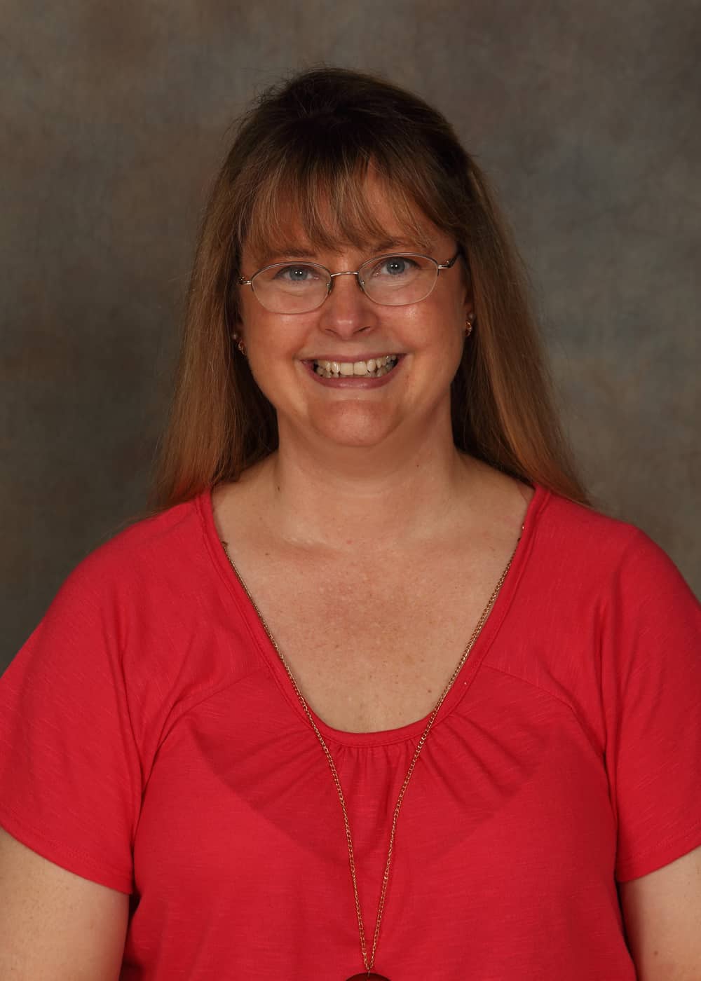 Photo of Lead Teacher Heidi Flynn. She is a Caucasian woman with long blonde hair, blue eyes and gold-rimmed glasses. She is smiling and wears a gold necklace and coral colored dress.