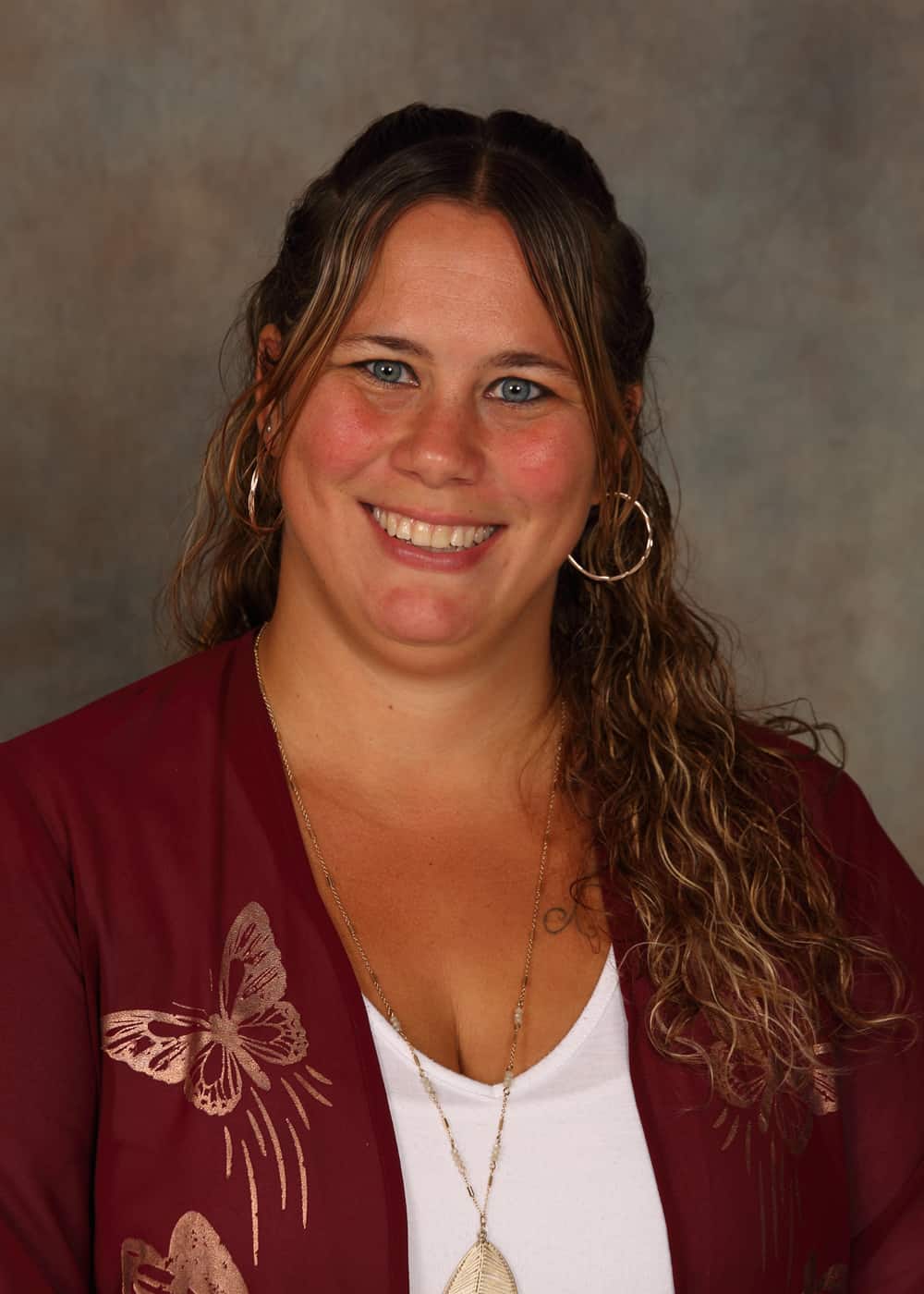 Photo of Lead Teacher Karrie Smith. She is a Caucasian woman with long brown hair, blue eyes, a bright smile and hoop earrings. She is wearing a white shirt and a dark red blouse with silver butterfly decorations.