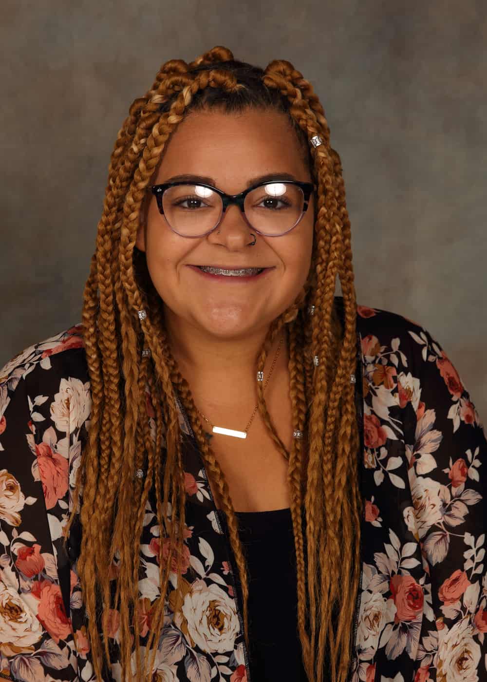Photo of Co-Teacher Marshell Davis. She is a smiling African-American woman with light brown braids, black-framed glasses, and a bright smile. She is wearing a black shirt and a black blouse with a white and pink floral pattern.