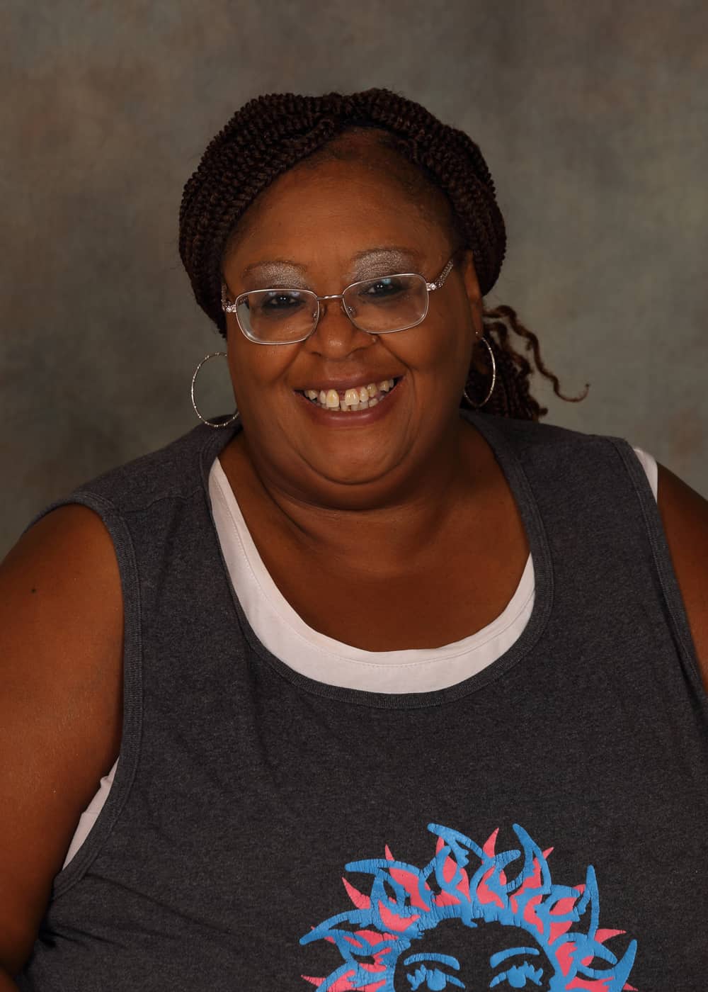 Photo of Program Director Priscilla McLin. She is an African-American woman with a bright smile, braided hair, hoop earrings and silver-rimmed glasses. She is wearing a dark gray shirt with a pink and blue picture of the Sun on it.