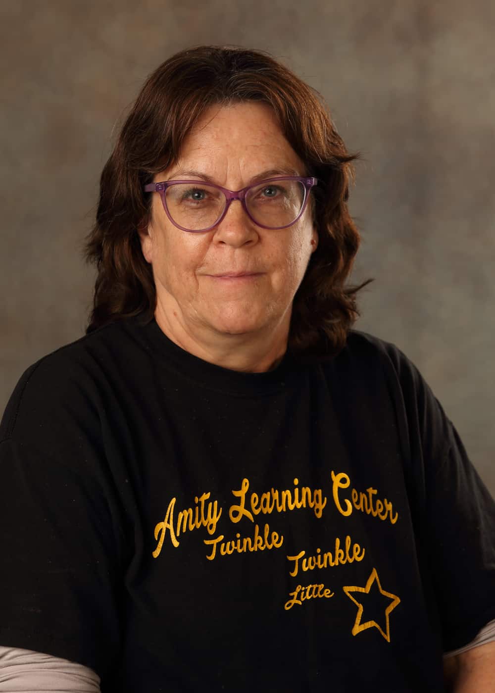 Photo of Kitchen Manager Rhonda Mashaw. She is a Caucasian woman with long dark brown hair, light blue eyes, and glasses with purple frames. She is wearing a black Amity Learning Center T-shirt with yellow lettering.