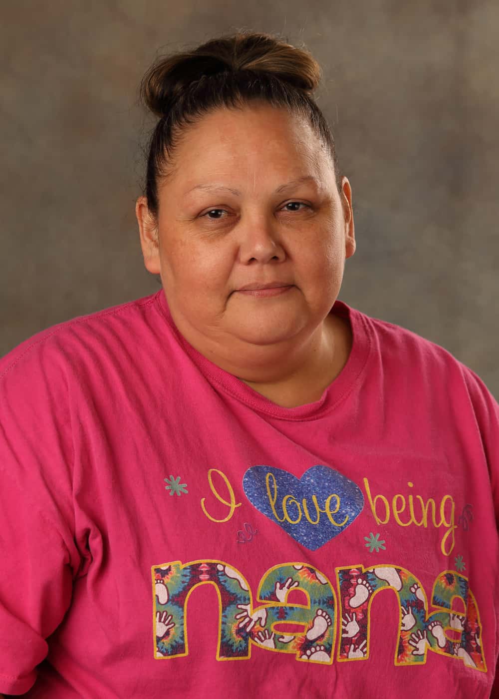 Photo of Kitchen Assistant Sara Callahan. She is a Caucasian woman with dark hair in a bun, and dark eyes. She is wearing a pink T-shirt that reads I love being Nana.