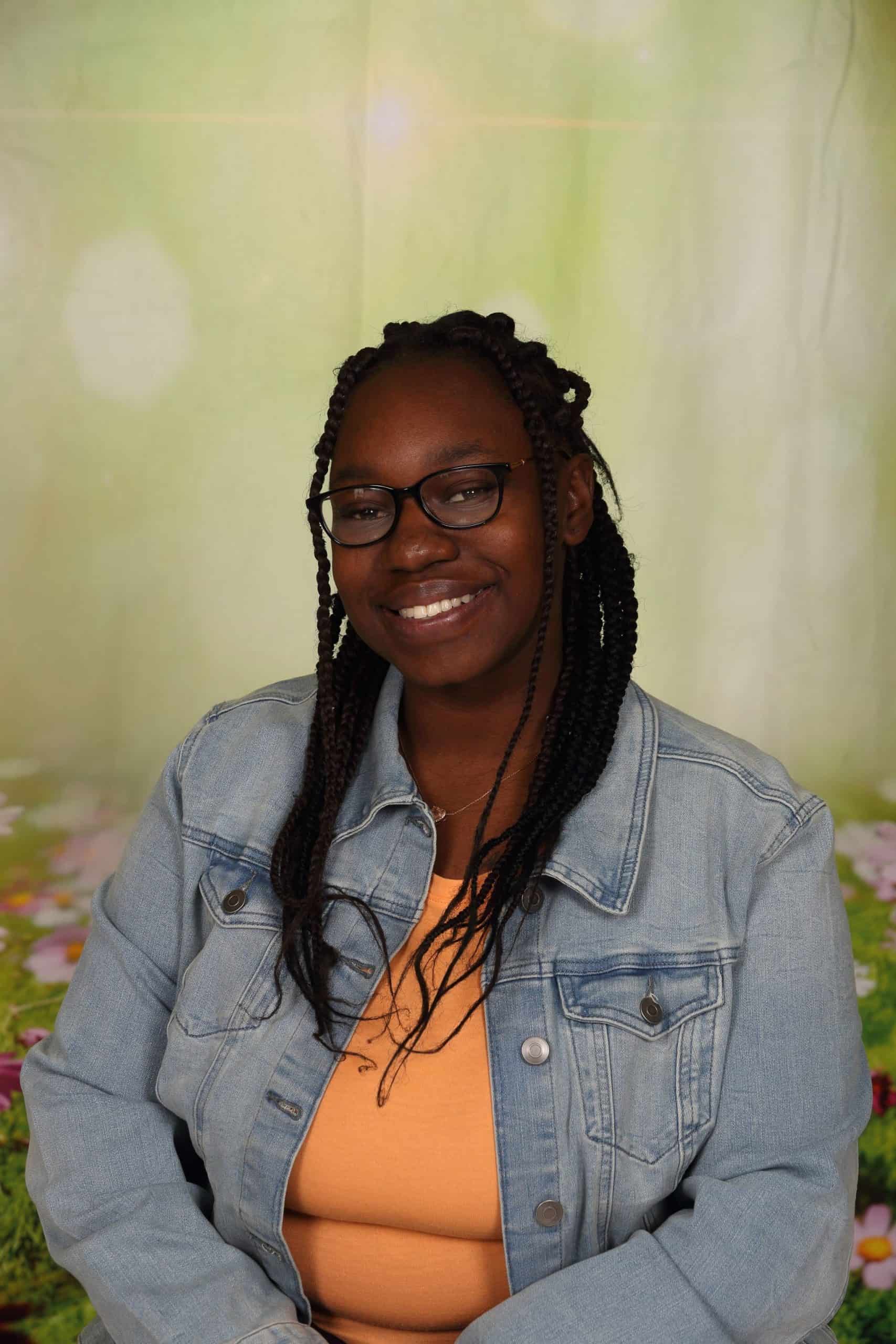Photo of Co-Teacher Audreyanna Thompson. She is a a young African-American woman with long hair. She is smiling andan orange shirt and denim jacket.
