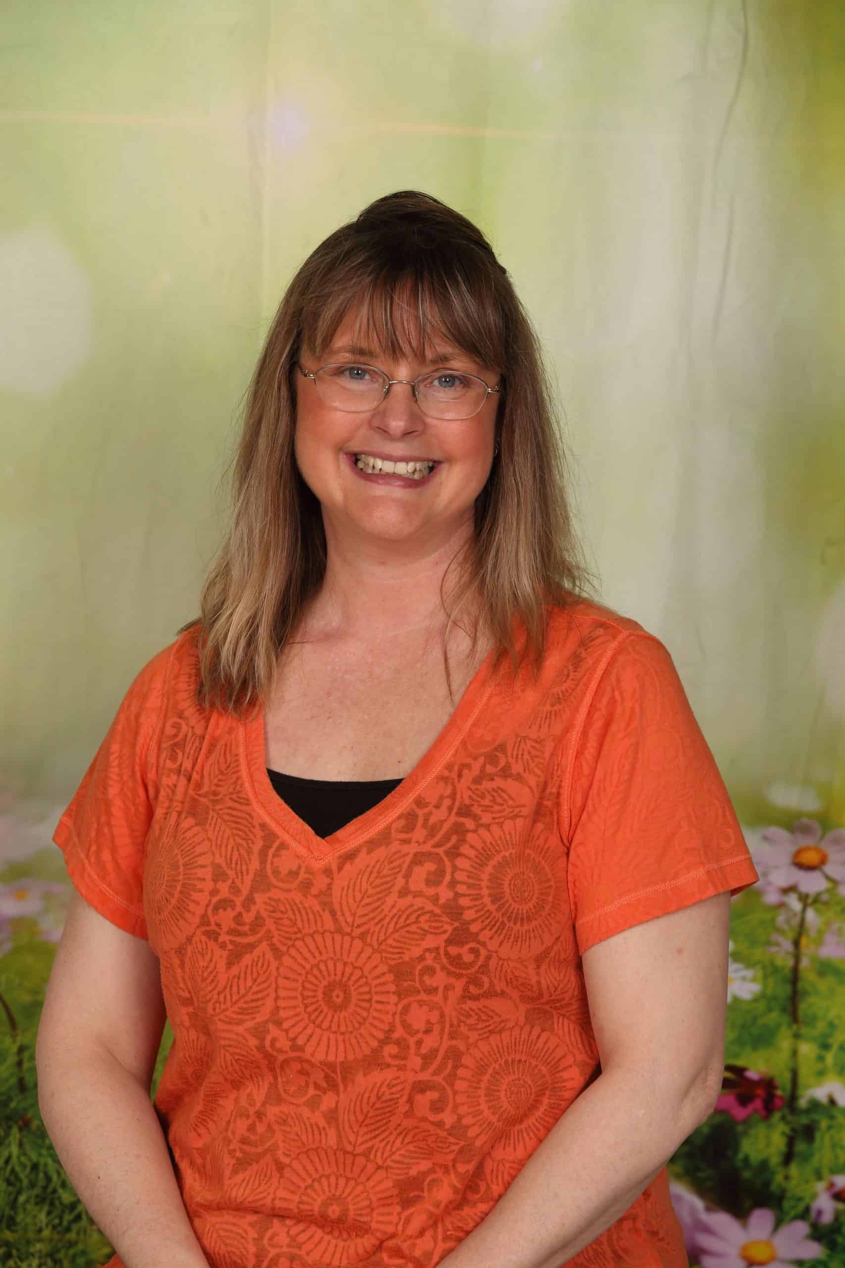 Photo of Lead Teacher Heidi Flynn. She is a Caucasian woman with long blonde hair, blue eyes and gold-rimmed glasses. She is smiling and wears a an orange shirt.